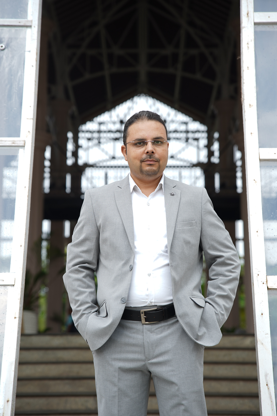 Dr Ashish Juneja, PhD Economics, standing in a light grey suit
